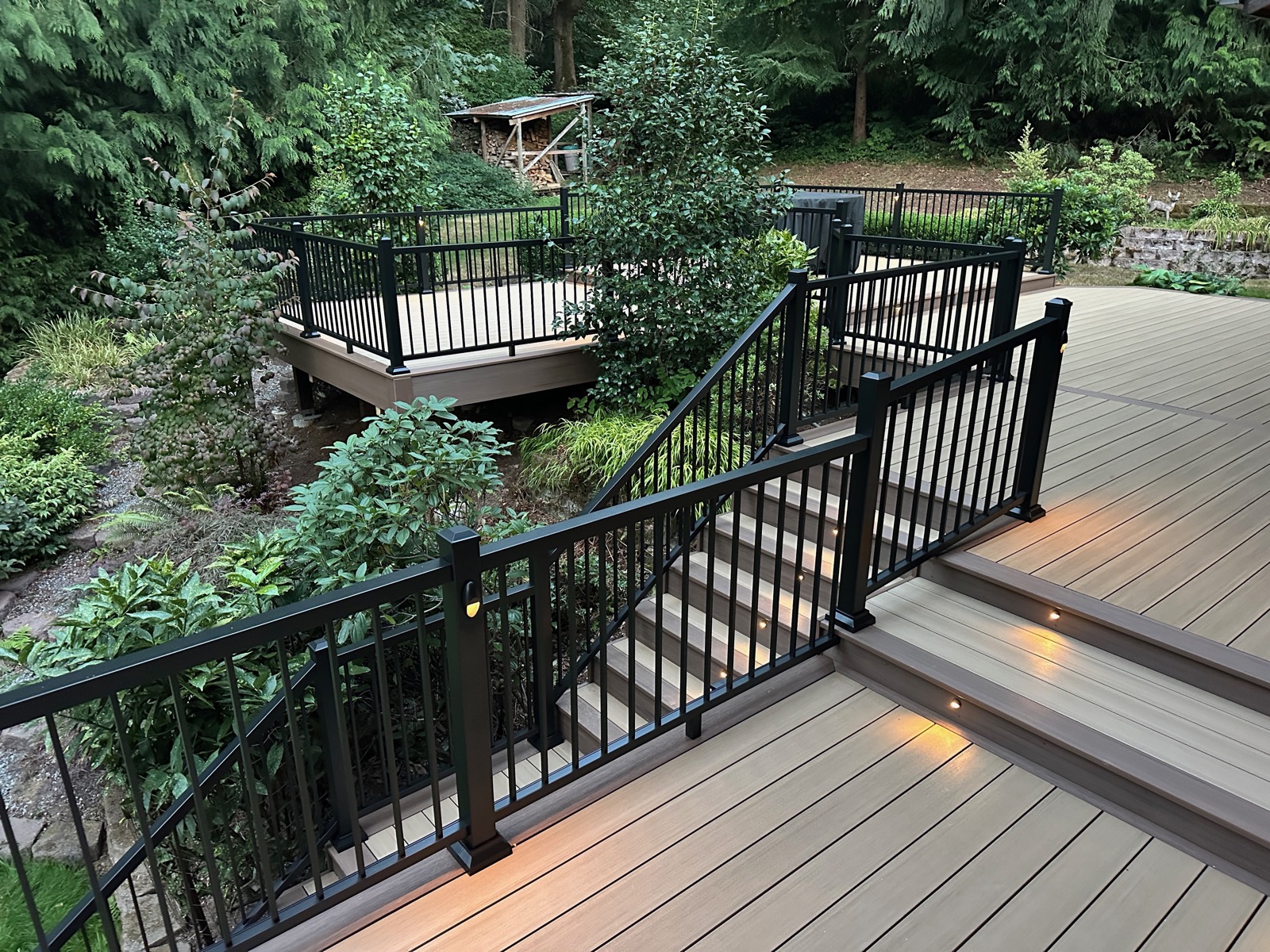 Composite stairs with black aluminum railing leading to a lower deck landing in a wooded yard