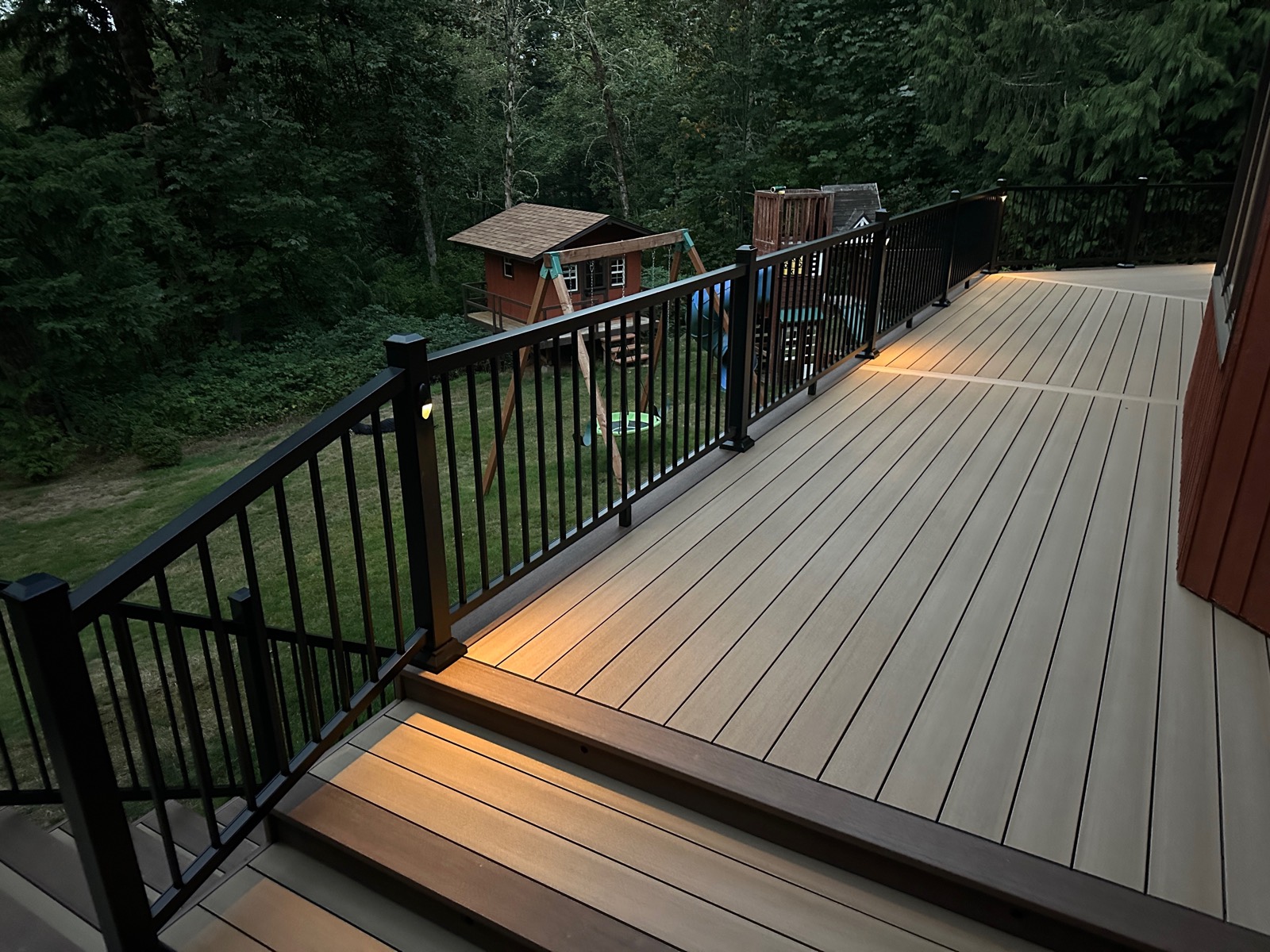 Composite deck with black railing and lit step overlooking a play area at dusk