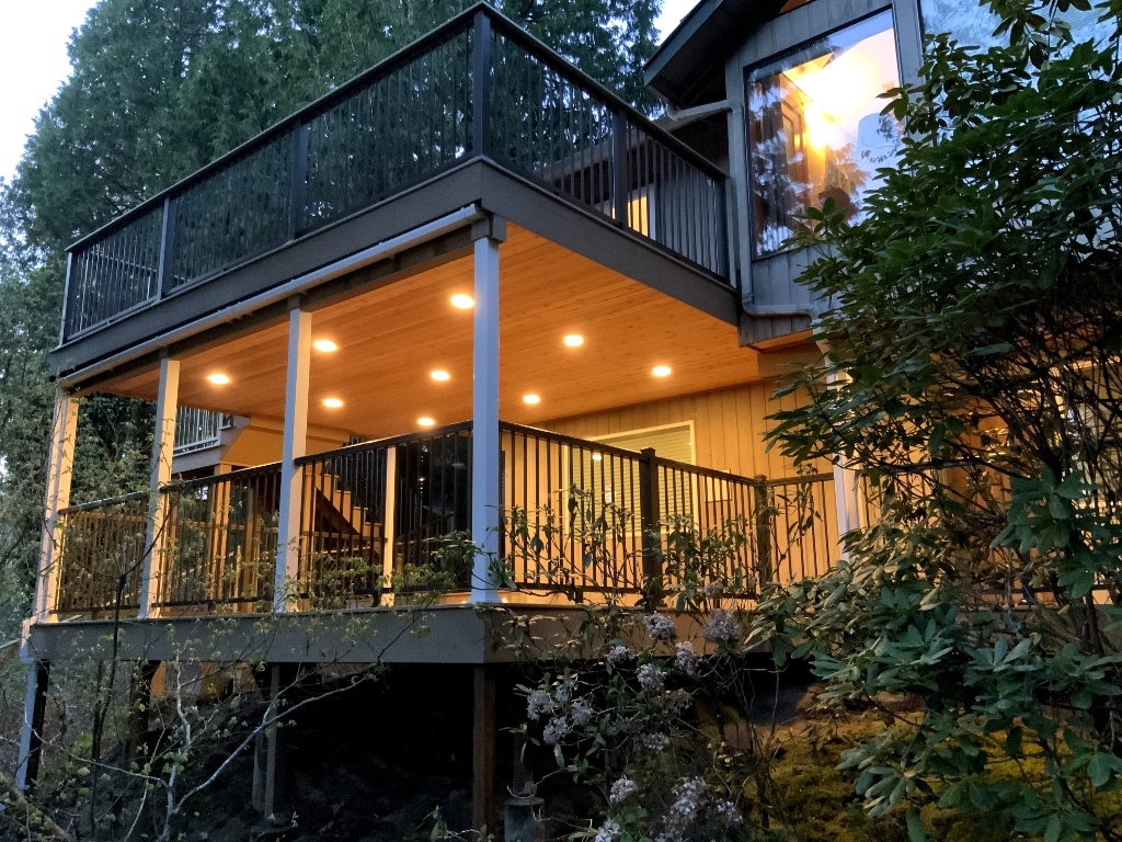 Elevated deck with glass-panel railing over a lit covered patio at dusk