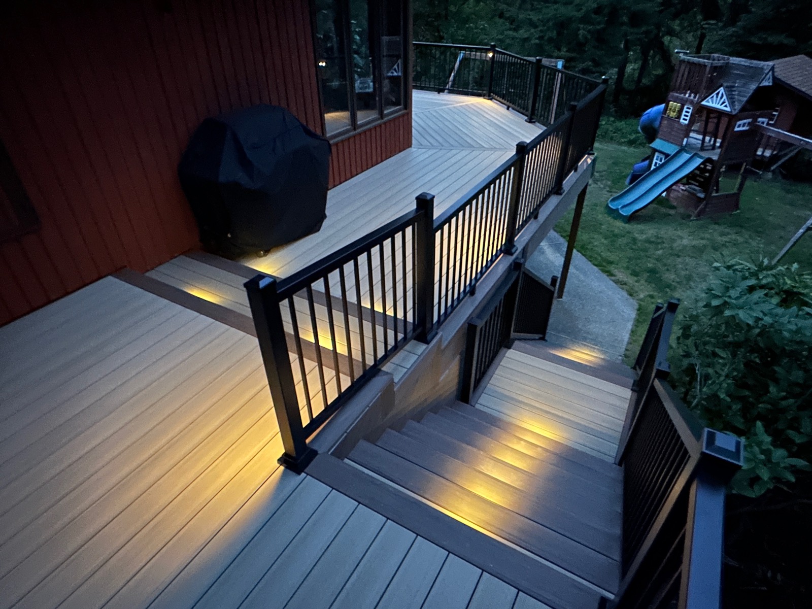 Composite deck stairs with black railing and riser lights beside a red home